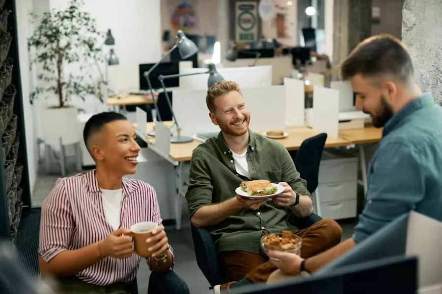 Employees on Lunch Break in Modern Office Workspace Three office employees enjoying a lunch break together in a modern workplace, smiling and talking while seated near desks. One holds a sandwich, another has a cup of coffee.