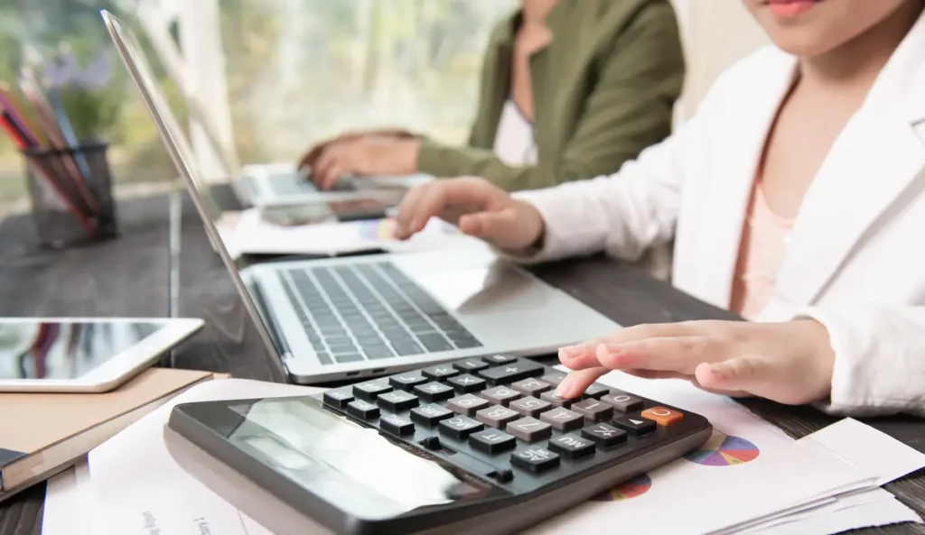 Employee calculating payroll hours using a calculator and laptop to ensure accurate wage and overtime calculations